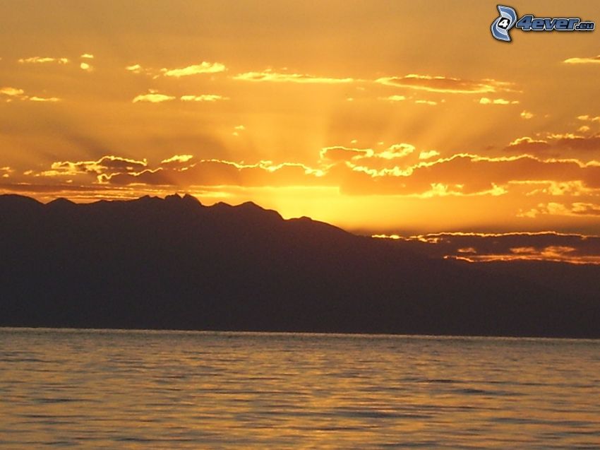 coucher du soleil derrière les montagnes, ciel orange, rayons du soleil, grand lac