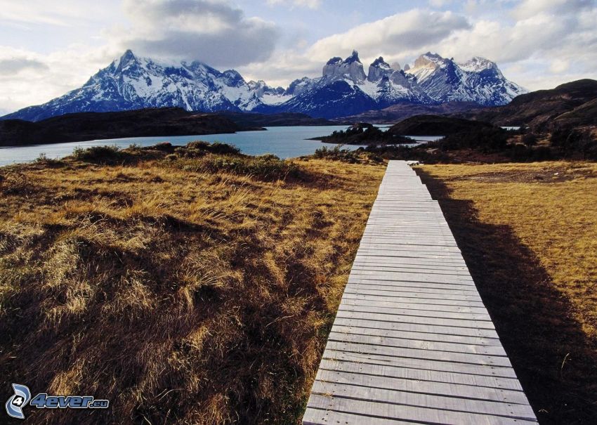 Torres del Paine, drevené mólo, zasnežené hory, rieka, pole